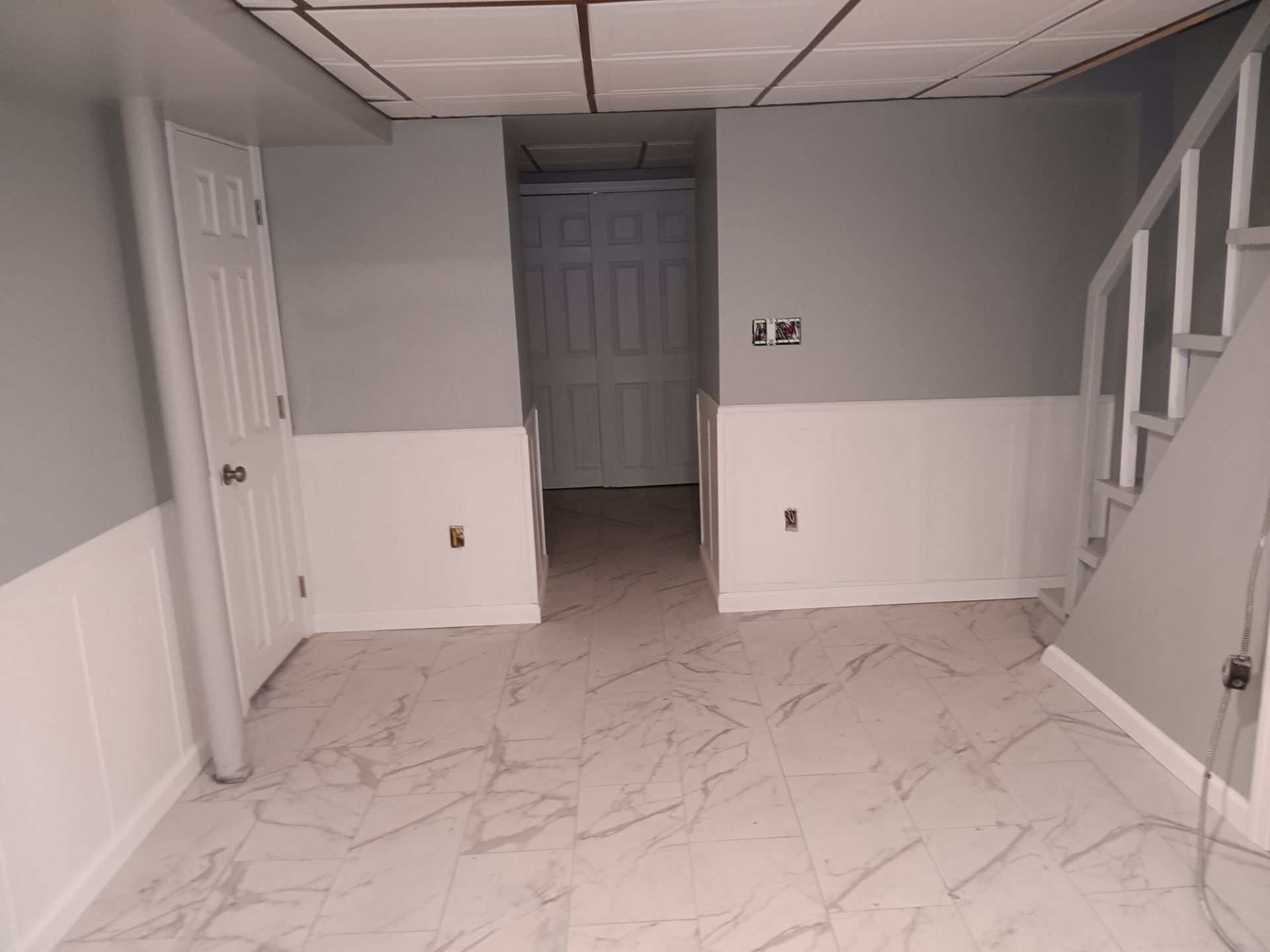 Empty basement room with white wainscoting, gray walls, tiled floor, and a staircase.