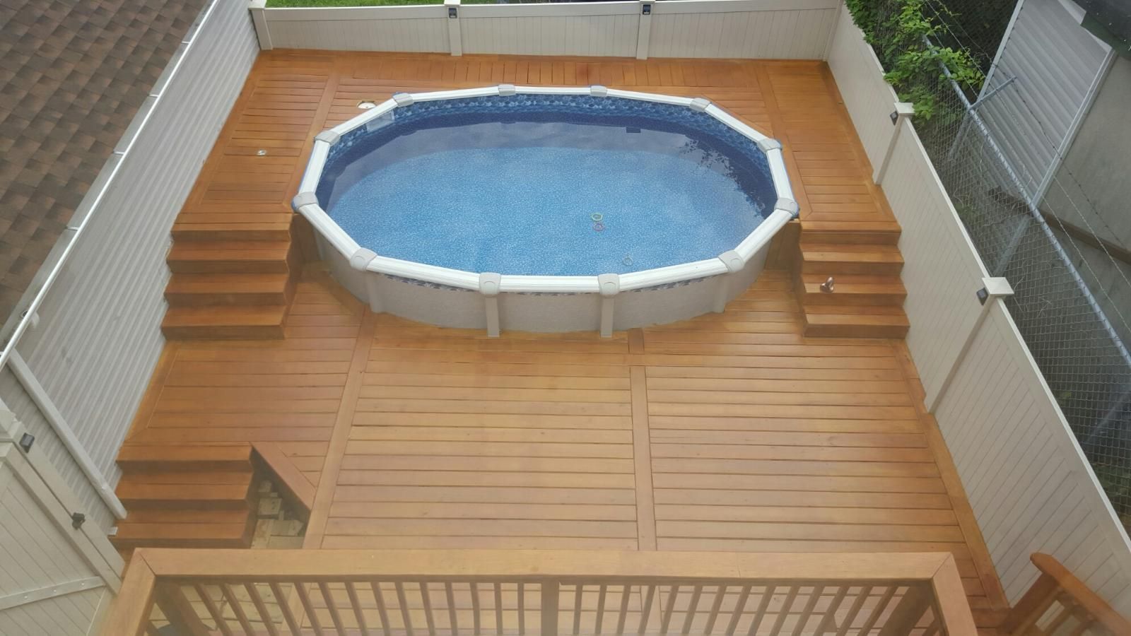 Above-ground pool on a wooden deck with steps on each side, surrounded by a white fence.