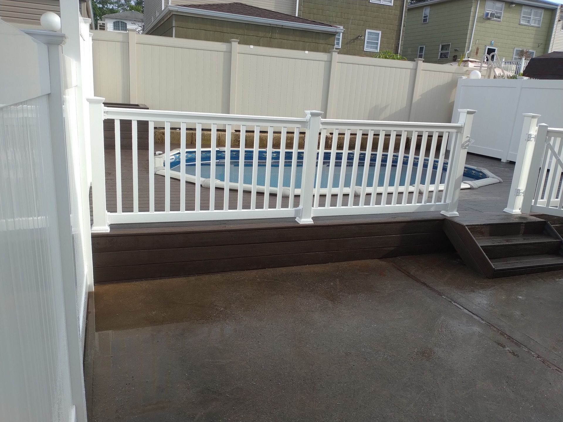 A fenced-in backyard with a pool, concrete patio, and steps. White fences, blue pool water, sunny day.