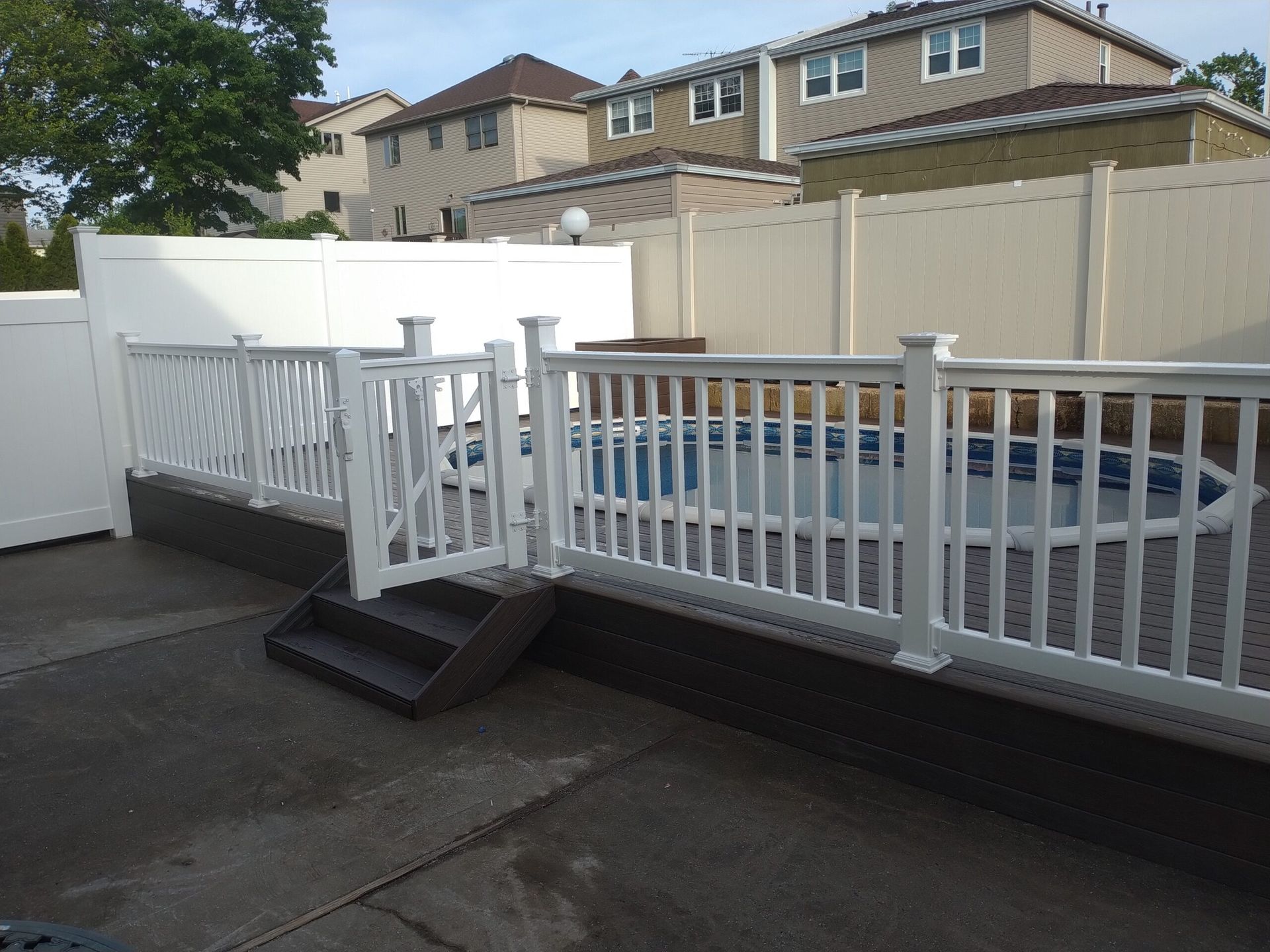 White fenced-in pool area with steps leading up to a deck, surrounded by residential houses.
