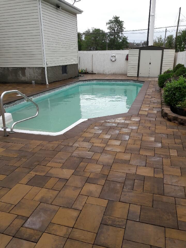 Pool with brown brick patio and a white fence, surrounded by a house and greenery.