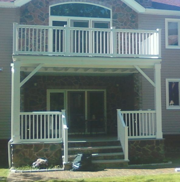 Two-story white deck with railing. Stone base, stairs lead to double doors. Building exterior in the background.