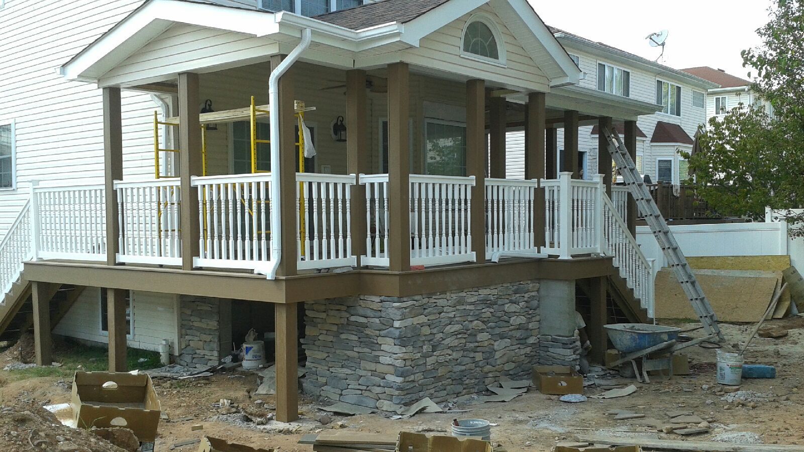 A porch with white railings, brown support beams, and stone foundation is under construction.