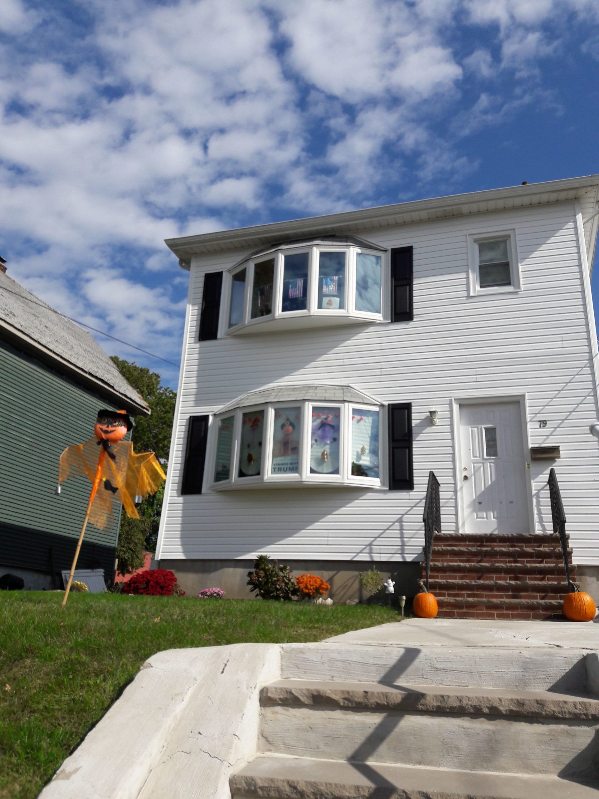 Two-story white house with bay windows, black shutters, and a scarecrow in the yard; sunny day.