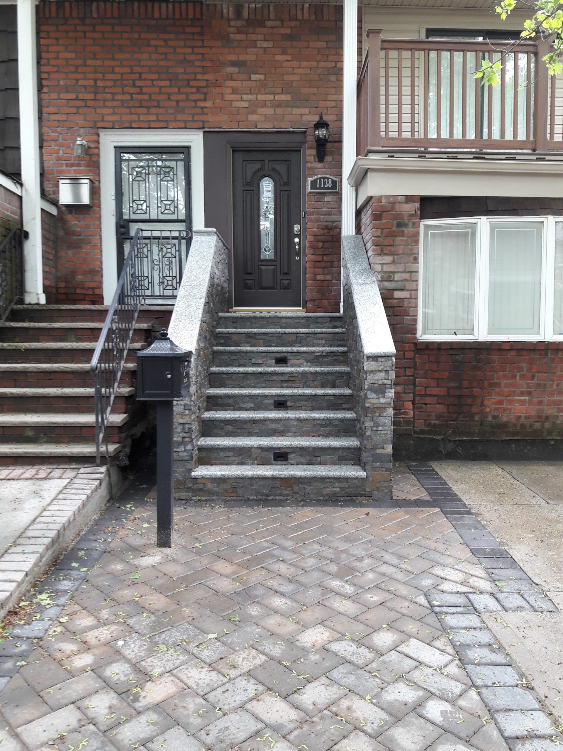 Brick home entrance with steps, decorative stone, and front door. Paved driveway and balcony.