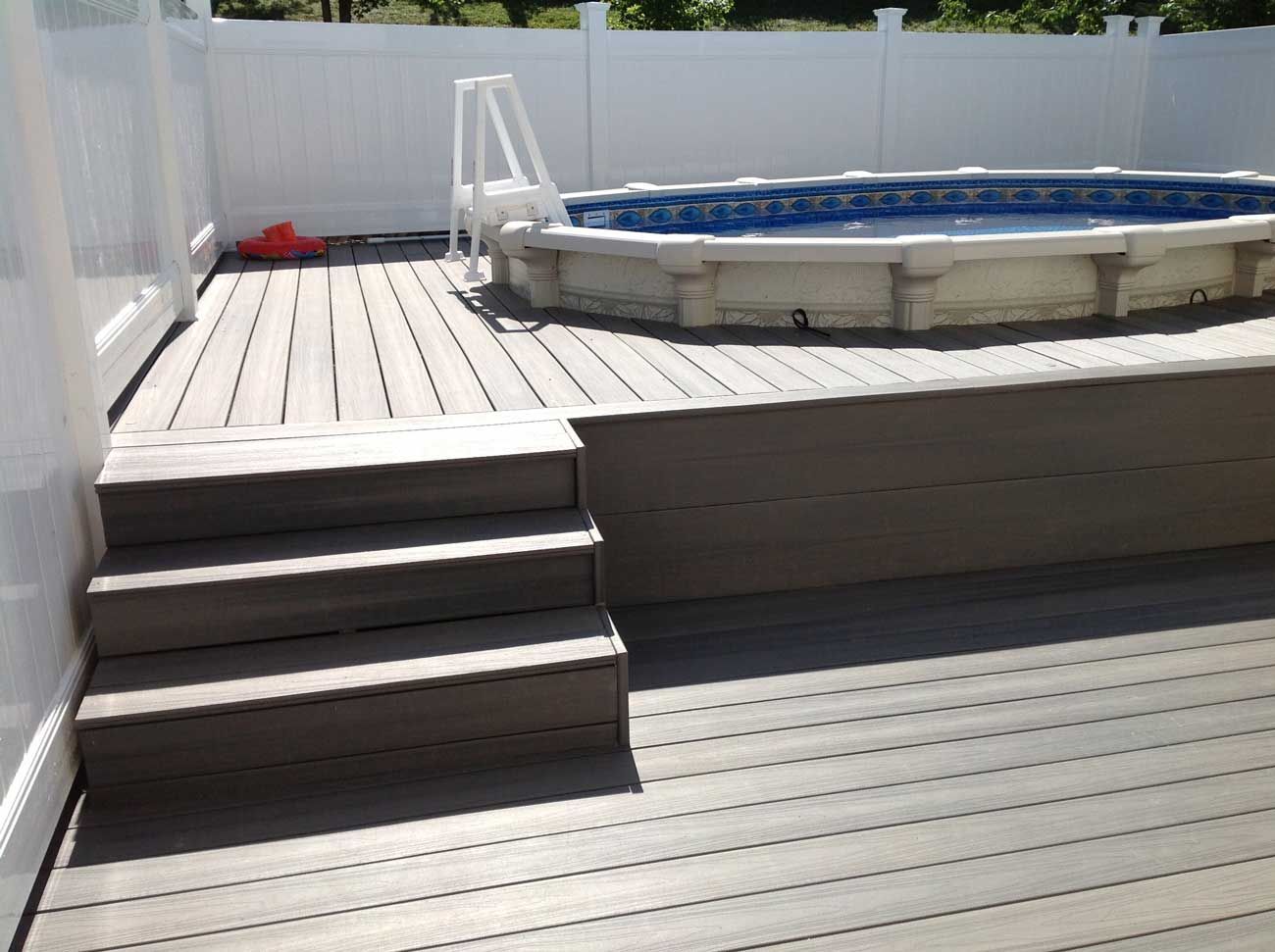 Above-ground pool on a deck with steps, enclosed by a white fence.