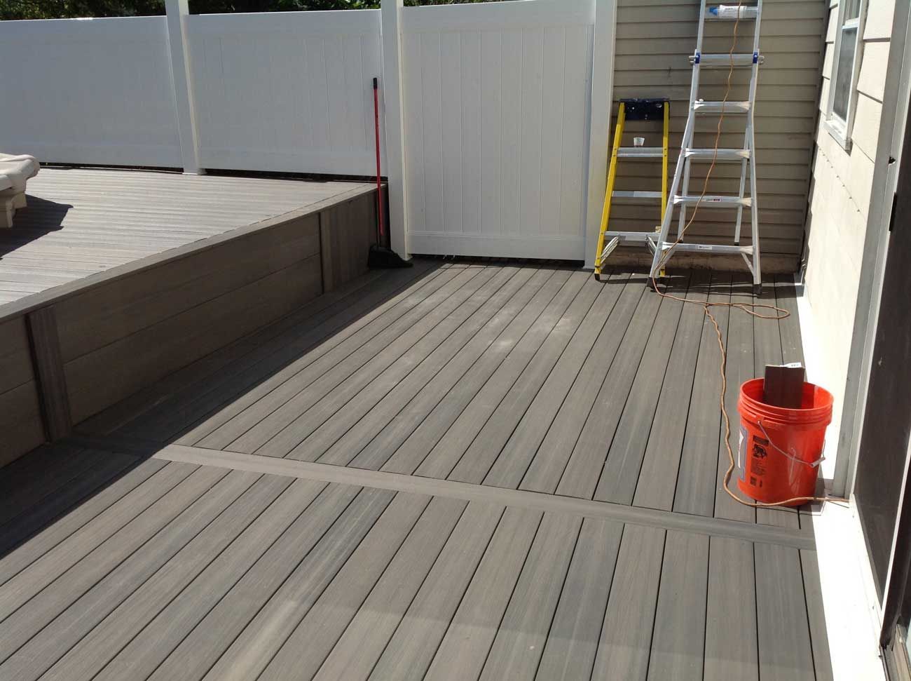 Composite deck with a raised edge, white fence, and a ladder leaning against a house. Orange bucket on the deck.
