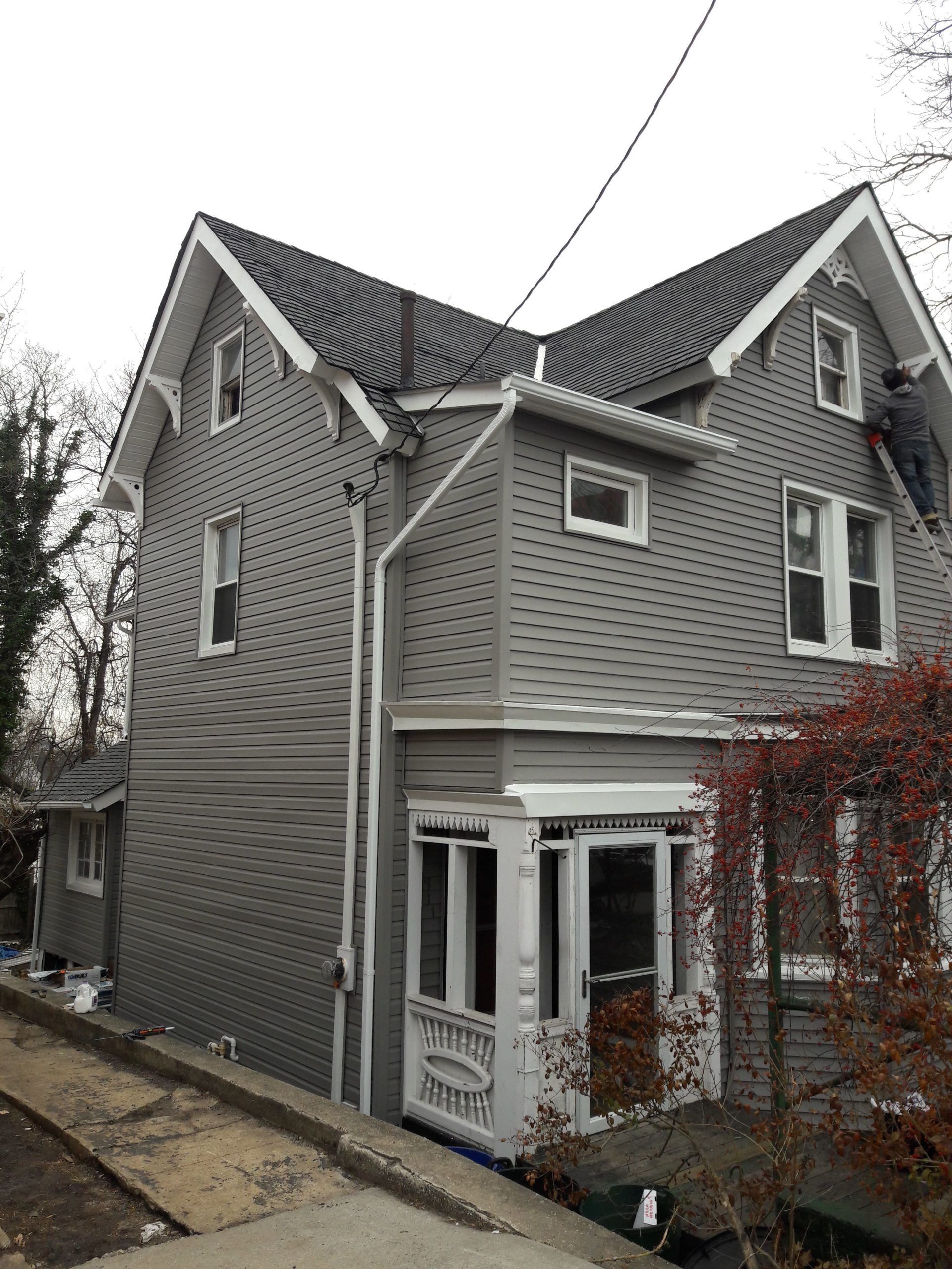 Two-story house with gray siding, white trim, and a dark roof. A person on a ladder is visible.