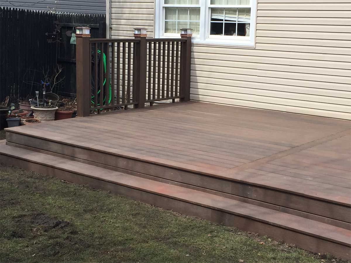 Wooden deck with steps, railing, and a hose, next to a vinyl-sided house on grass.