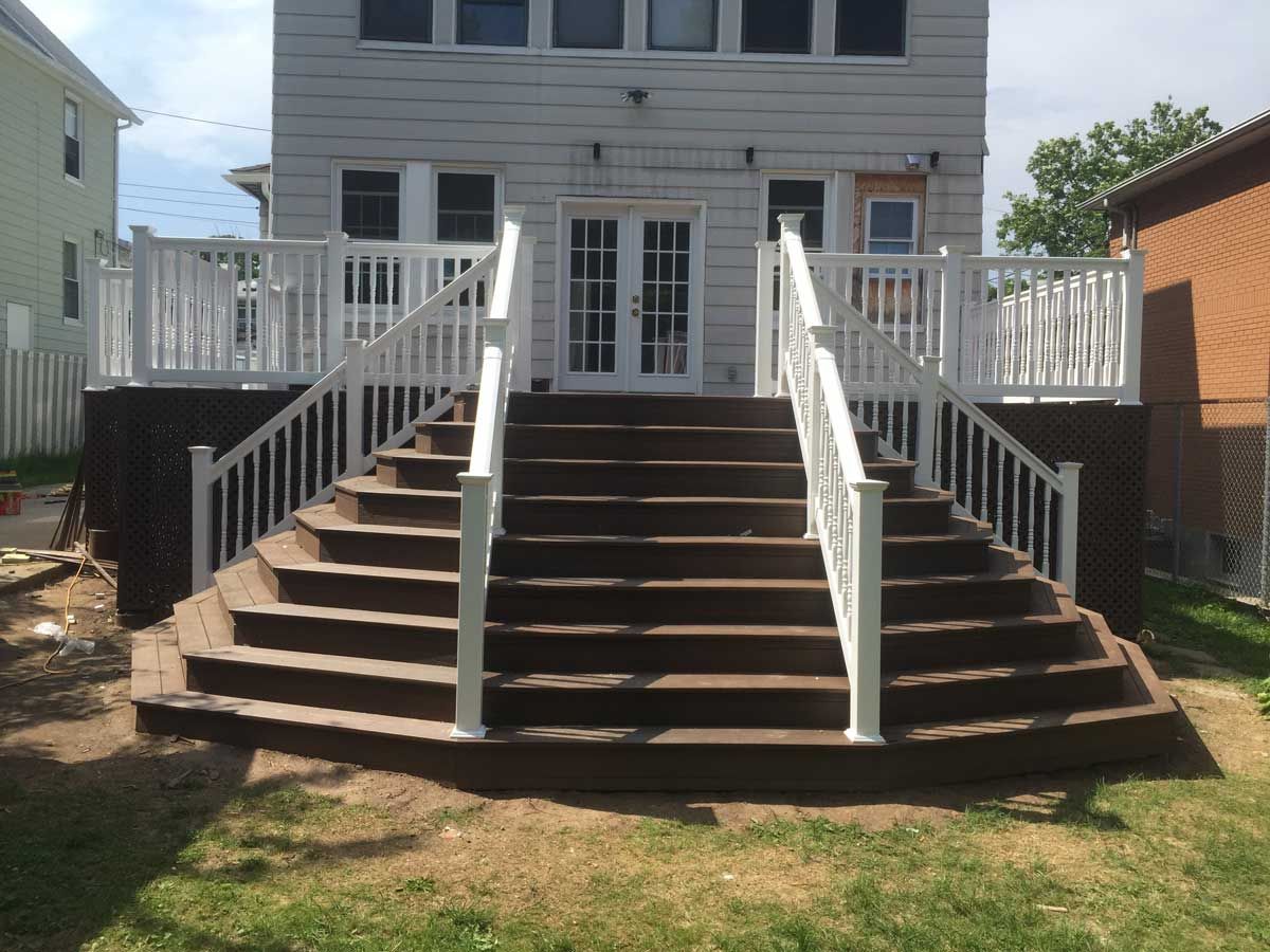 Brown wooden deck with white railing, leading to a house with double doors.