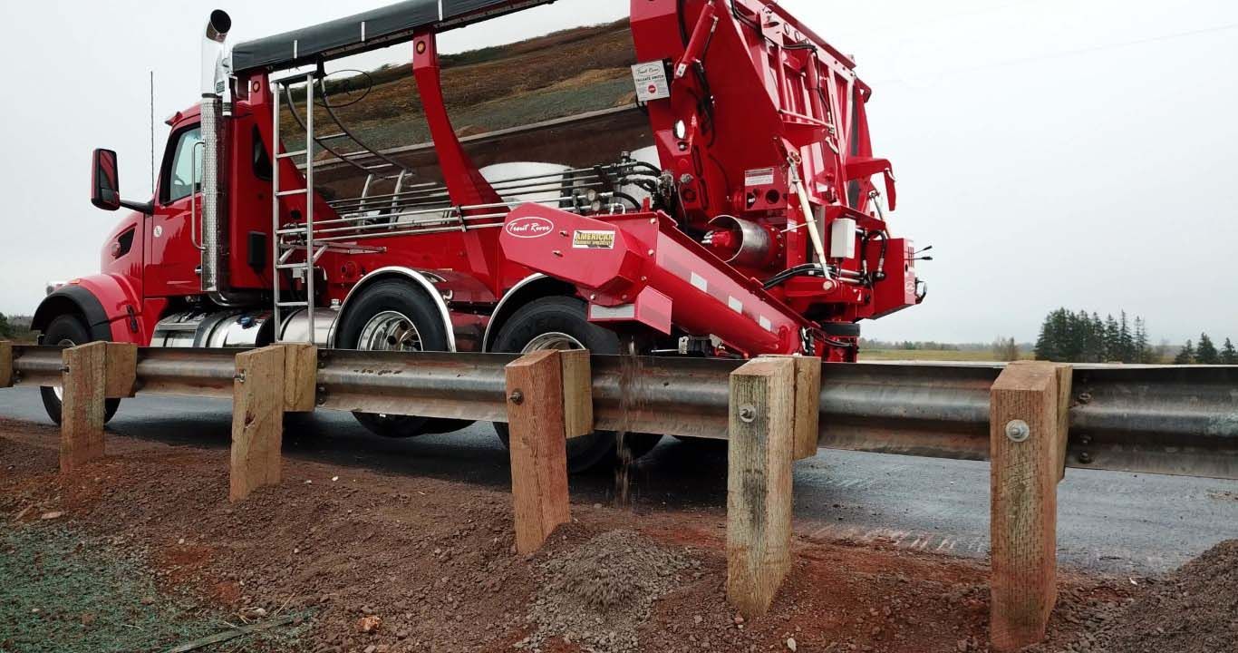 Trout River Industries truck-mounted live bottom auger dispensing gravel over a bridge guard rail view from the side
