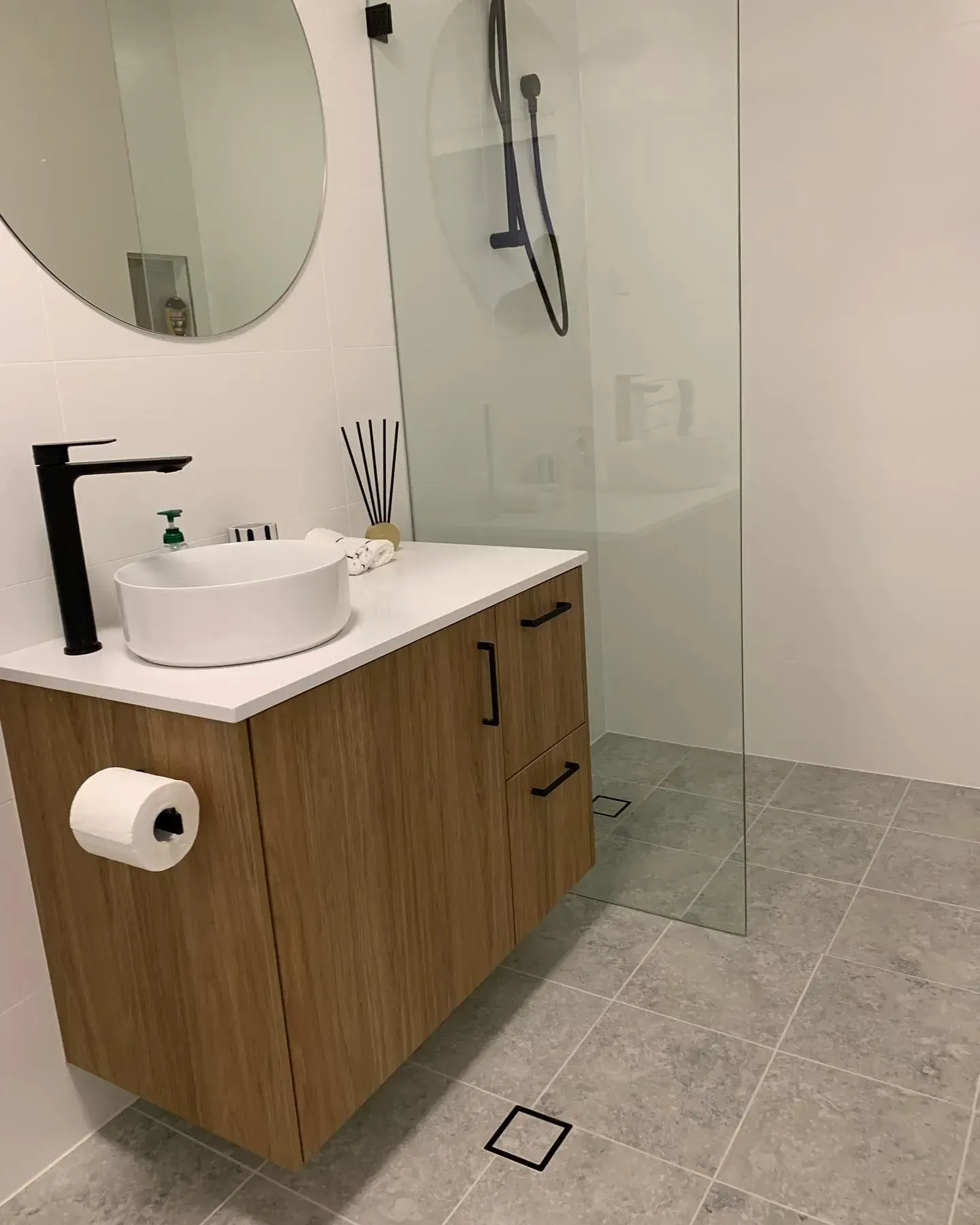 Bathroom With Wooden Vanity, Round Mirror, Glass Shower, and Gray Tiled Floor — The Kitchen Centre In Tuggerah, NSW