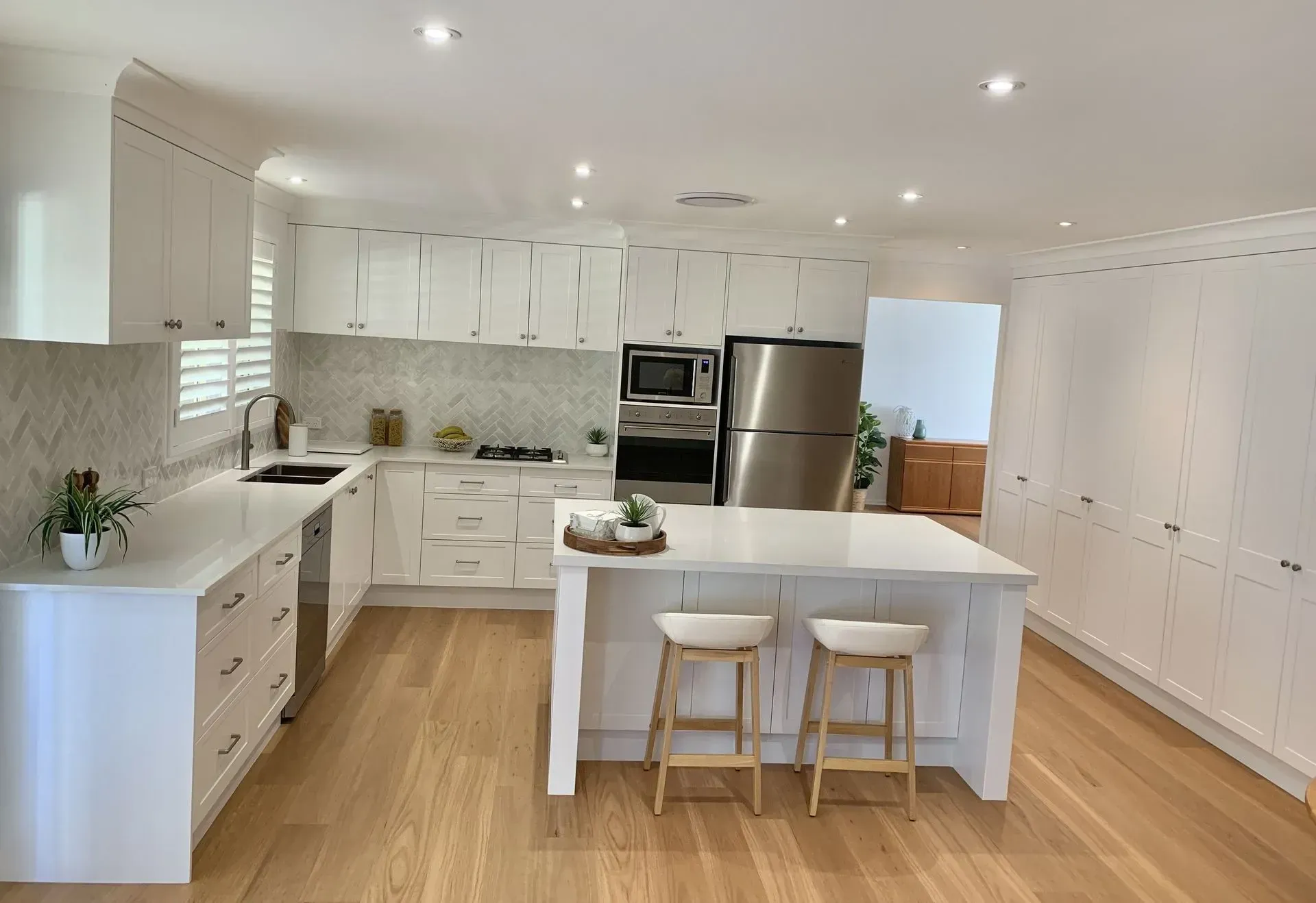 White Kitchen With Island, Wood Floor, Stainless Steel Fridge, and Two Bar Stools — The Kitchen Centre In Tuggerah, NSW
