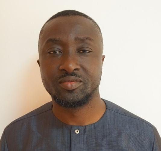 Man with dark skin, wearing a blue shirt, looking directly at the camera against a white background.
