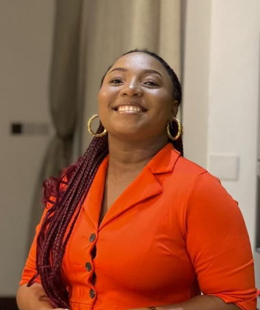 Woman in orange dress smiles, wearing gold hoop earrings. Braided hair with red highlights. Indoor setting.