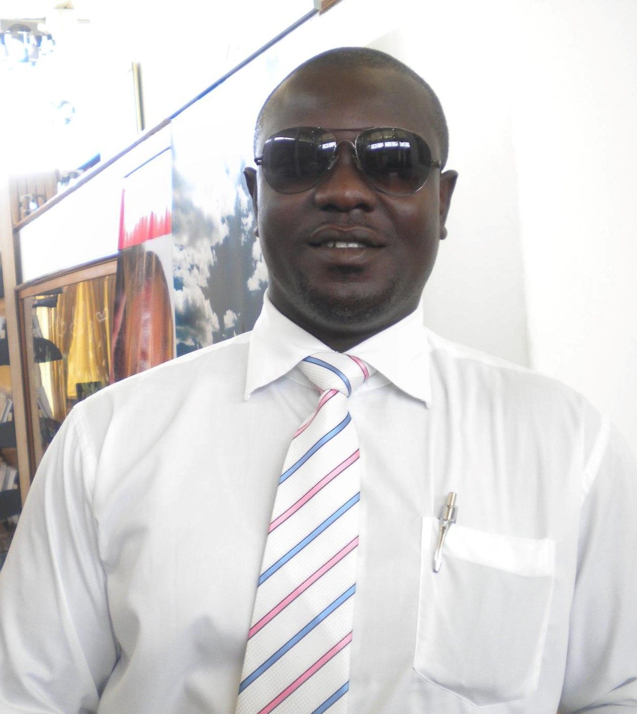 Man in sunglasses, white shirt, striped tie, and pocket pen, indoors, facing the camera.