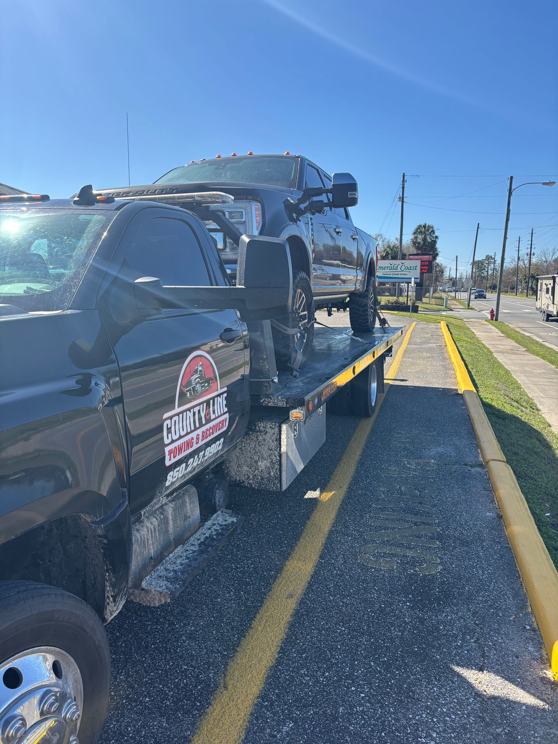 A black tow truck carrying a black truck on a sunny day next to a road.