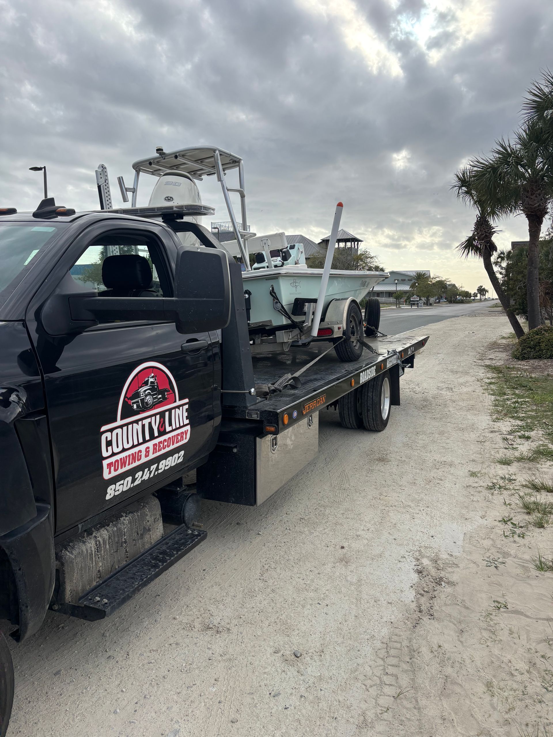 Black tow truck transporting a boat on a trailer along a sandy road. Cloudy sky.
