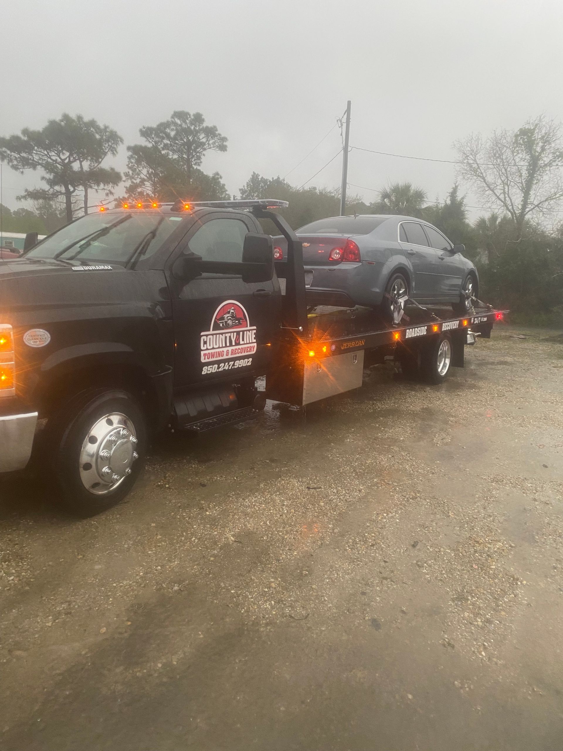A black tow truck with a gray car on its flatbed, on a wet asphalt road under a cloudy sky.