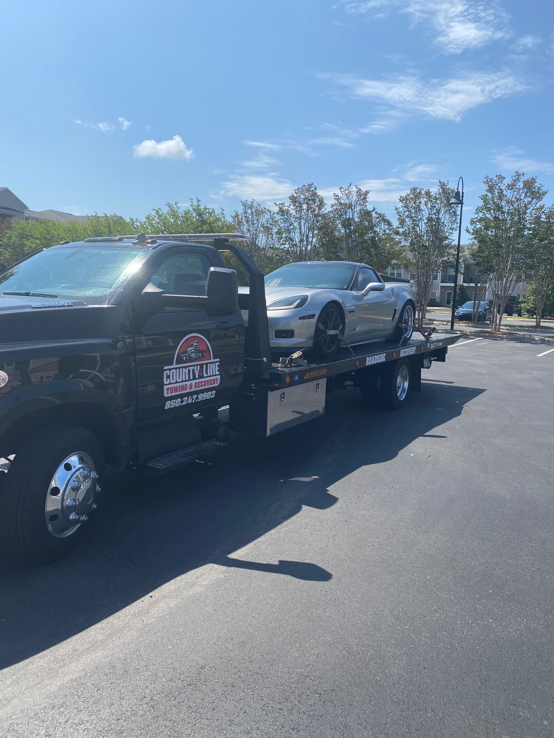 Silver Corvette on a black tow truck in a parking lot on a sunny day.