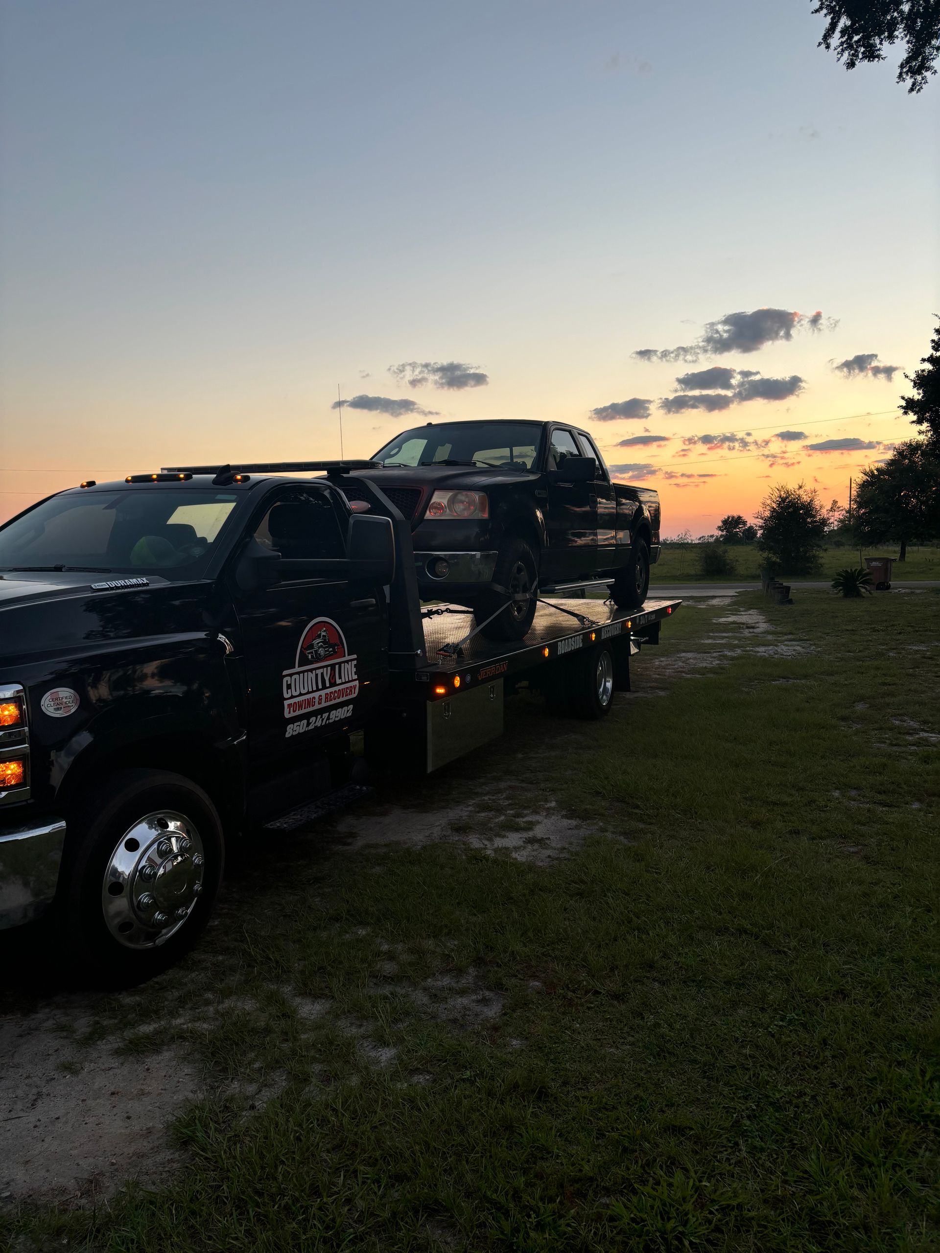Tow truck with a black pickup truck on its flatbed at sunset.