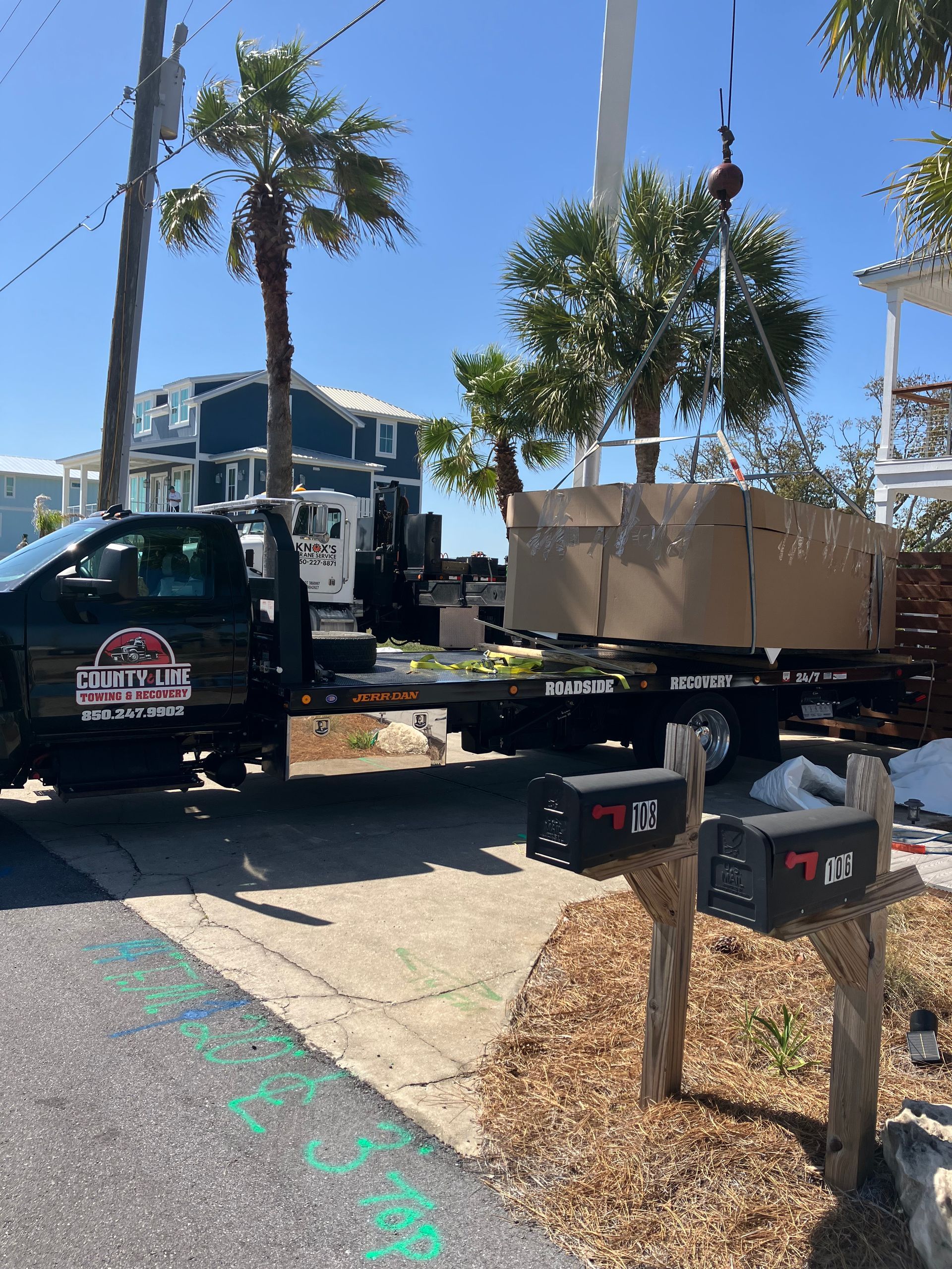 A truck with a box on a flatbed being lifted by a crane near mailboxes and houses on a sunny day.