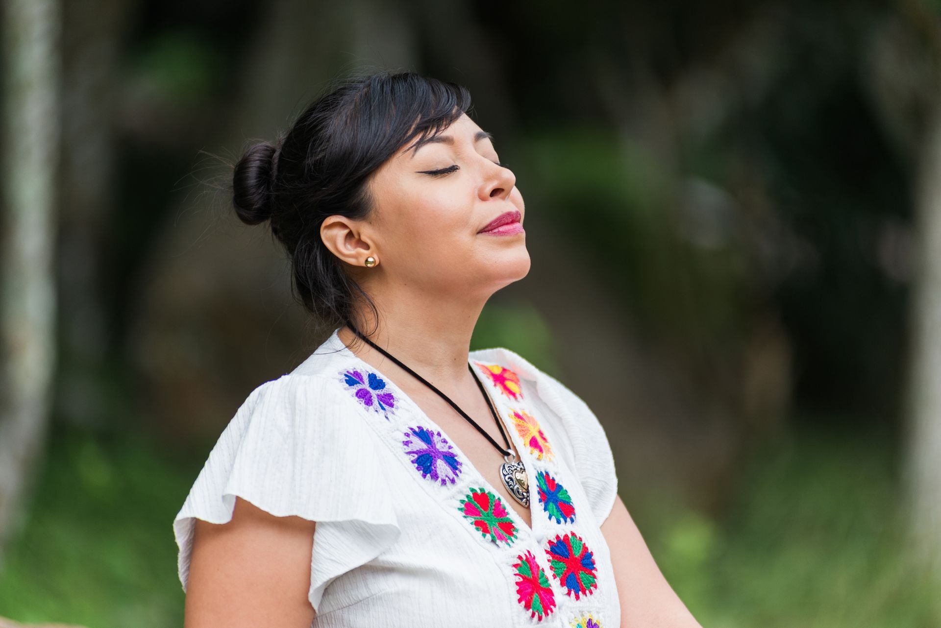 A person with hair in a bun wearing an embroidered white top, eyes closed in a calm expression outdoors.