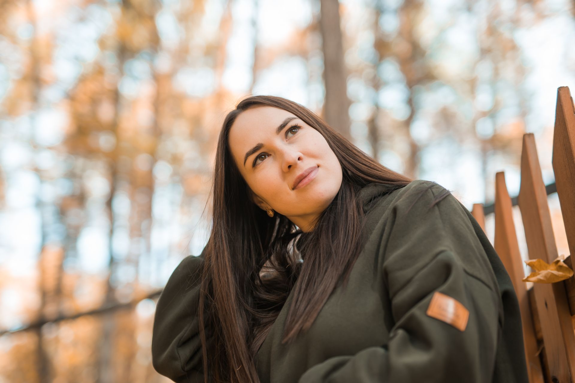 A person with long dark hair, wearing a green hooded sweatshirt, looks upward while leaning against a wooden fence.
