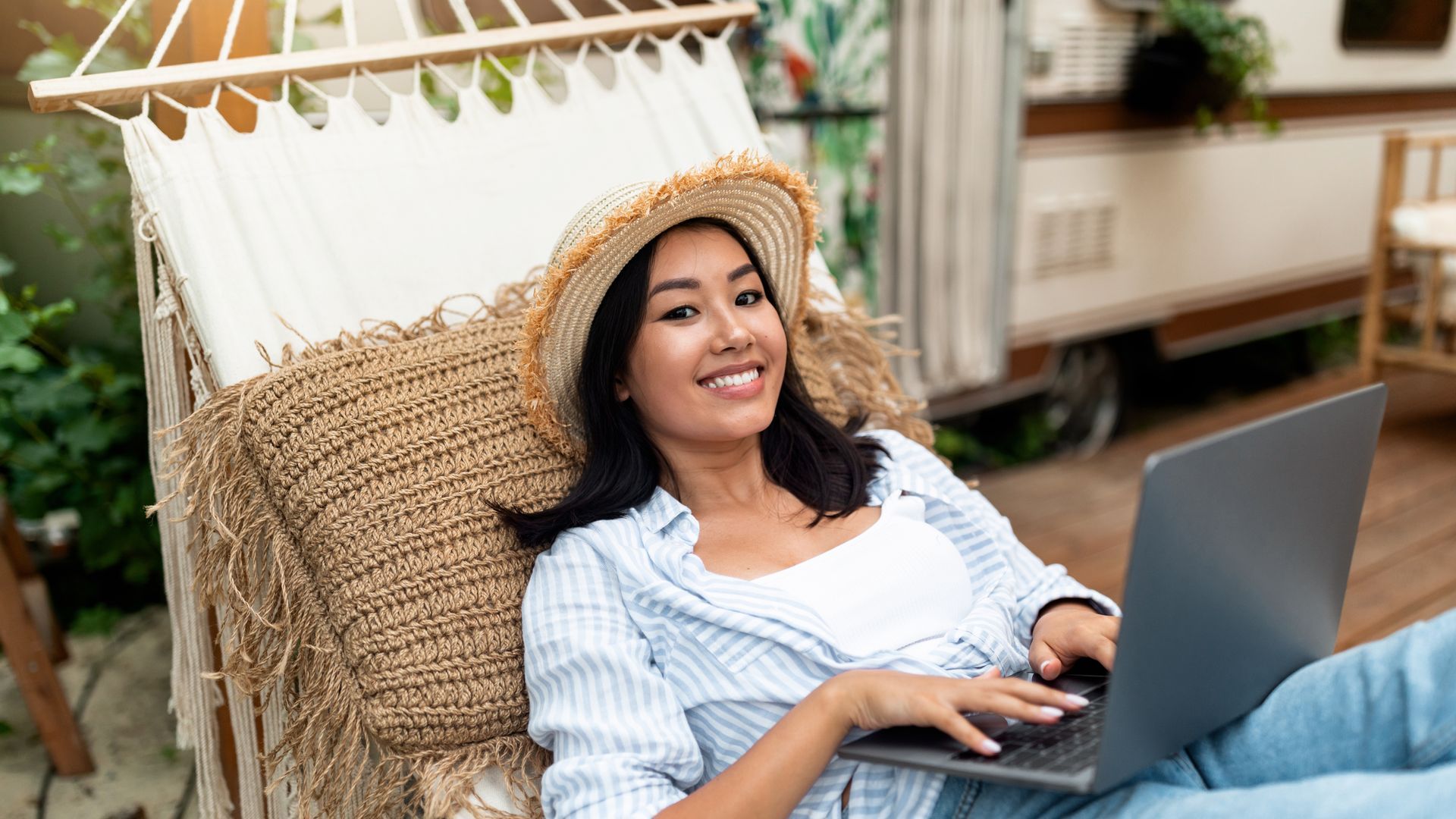 A smiling person wearing a straw hat rests in a hammock, working on a laptop with a camper in the background.