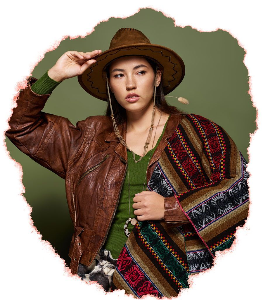 A person wears a brown leather jacket, green shirt, and patterned poncho with a fedora, posed against a green background.