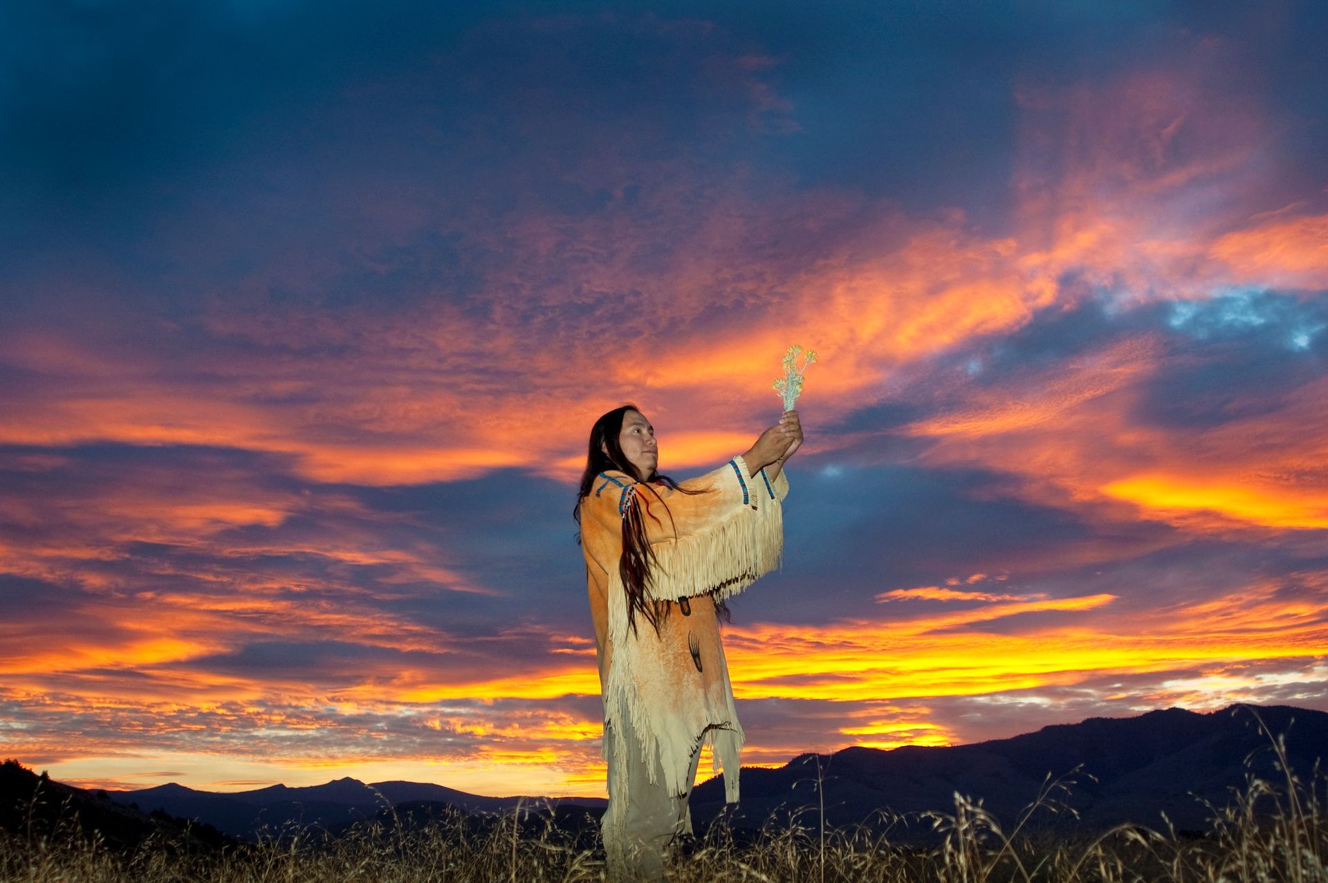 A person in traditional tan fringed clothing holds a feather aloft in a field against a vibrant, orange-streaked sunset.