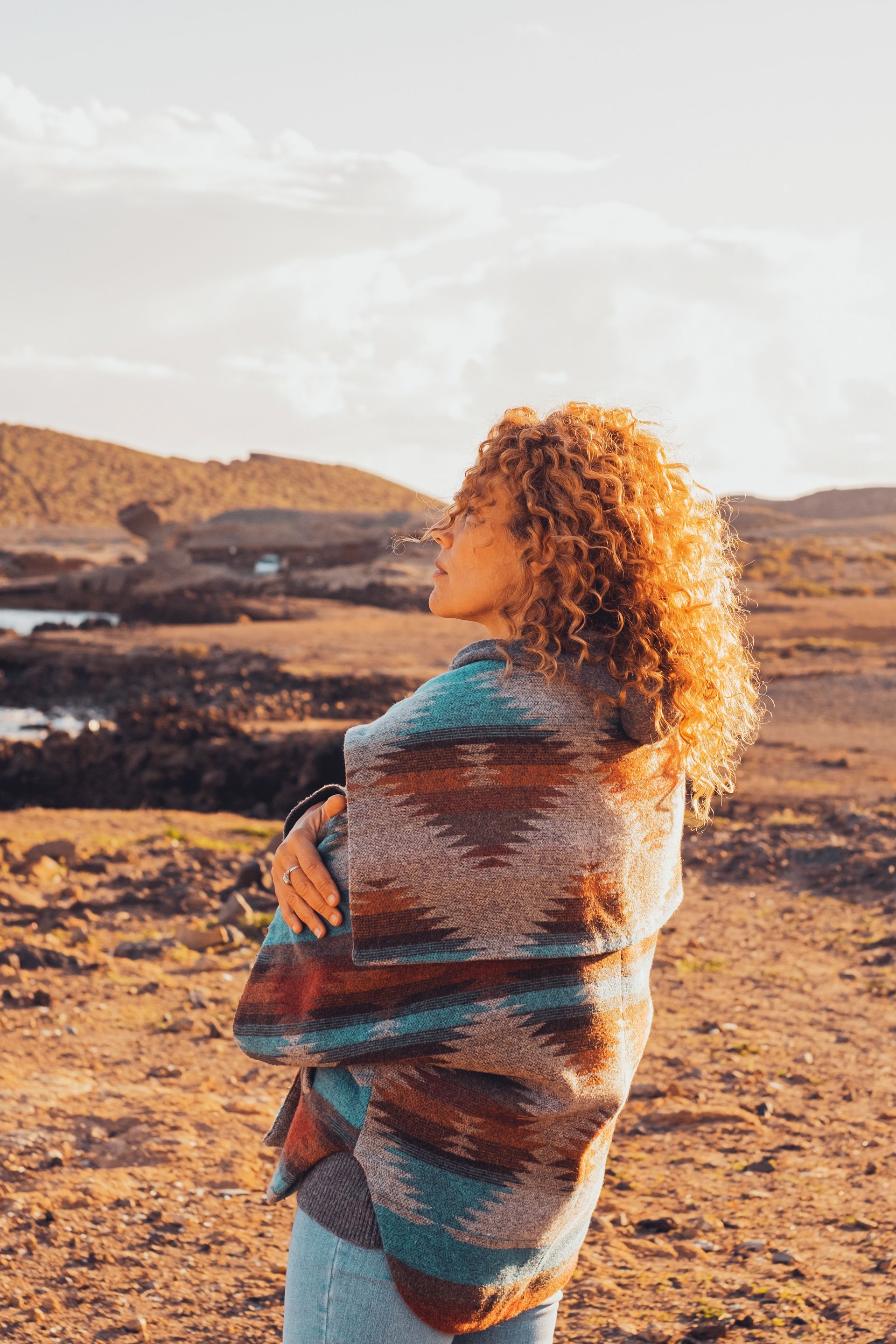 A curly-haired person wrapped in a patterned, earth-toned blanket stands in a sunlit, rocky landscape.