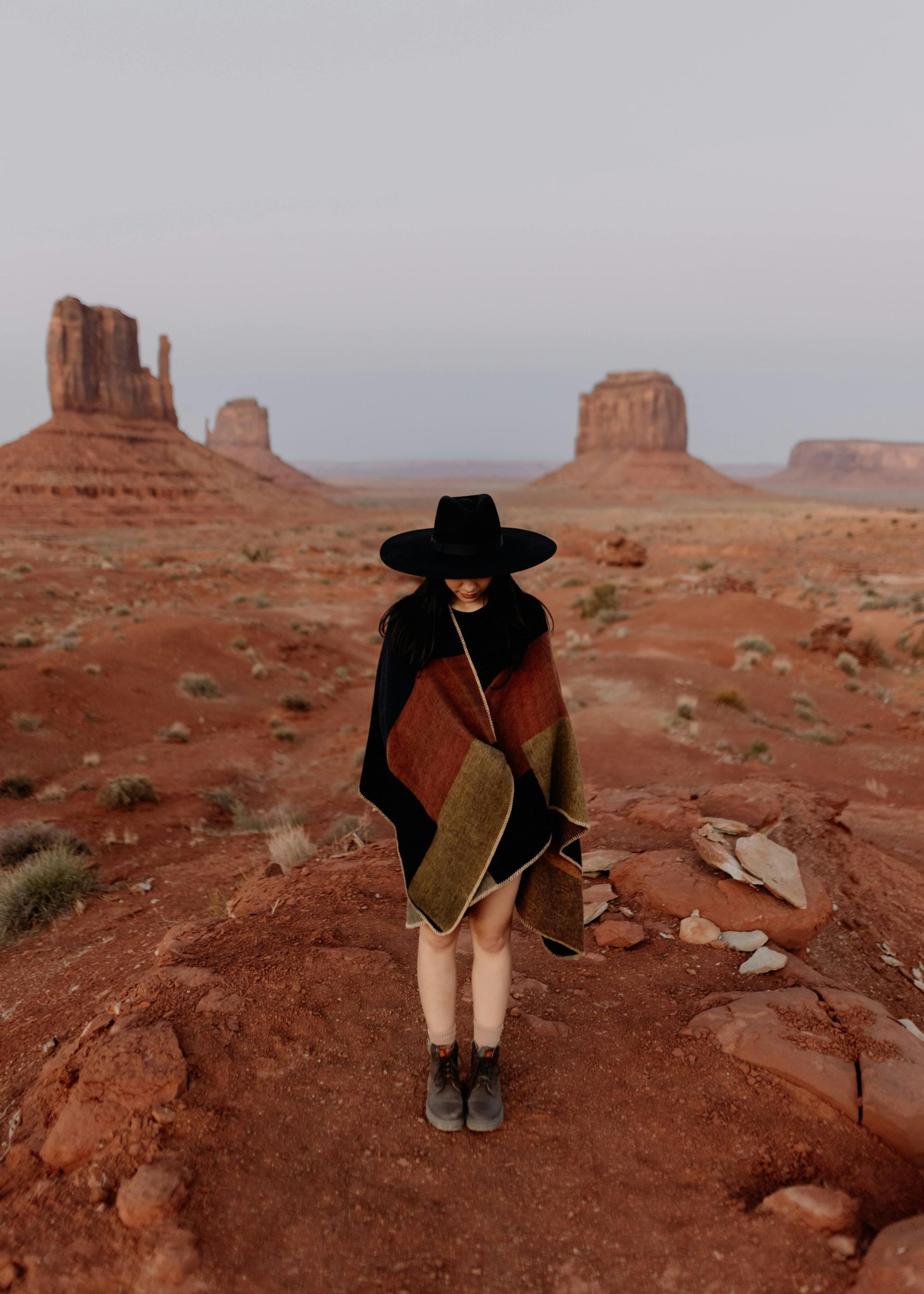 A person in a black hat and poncho stands on red desert terrain with Monument Valley buttes in the background.
