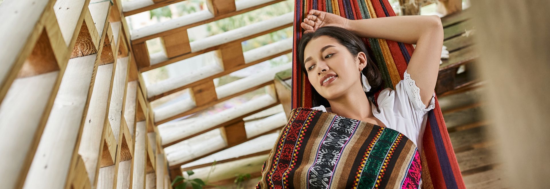 A person with a neutral expression rests in a colorful, striped hammock inside a structure made of light-colored wood poles.