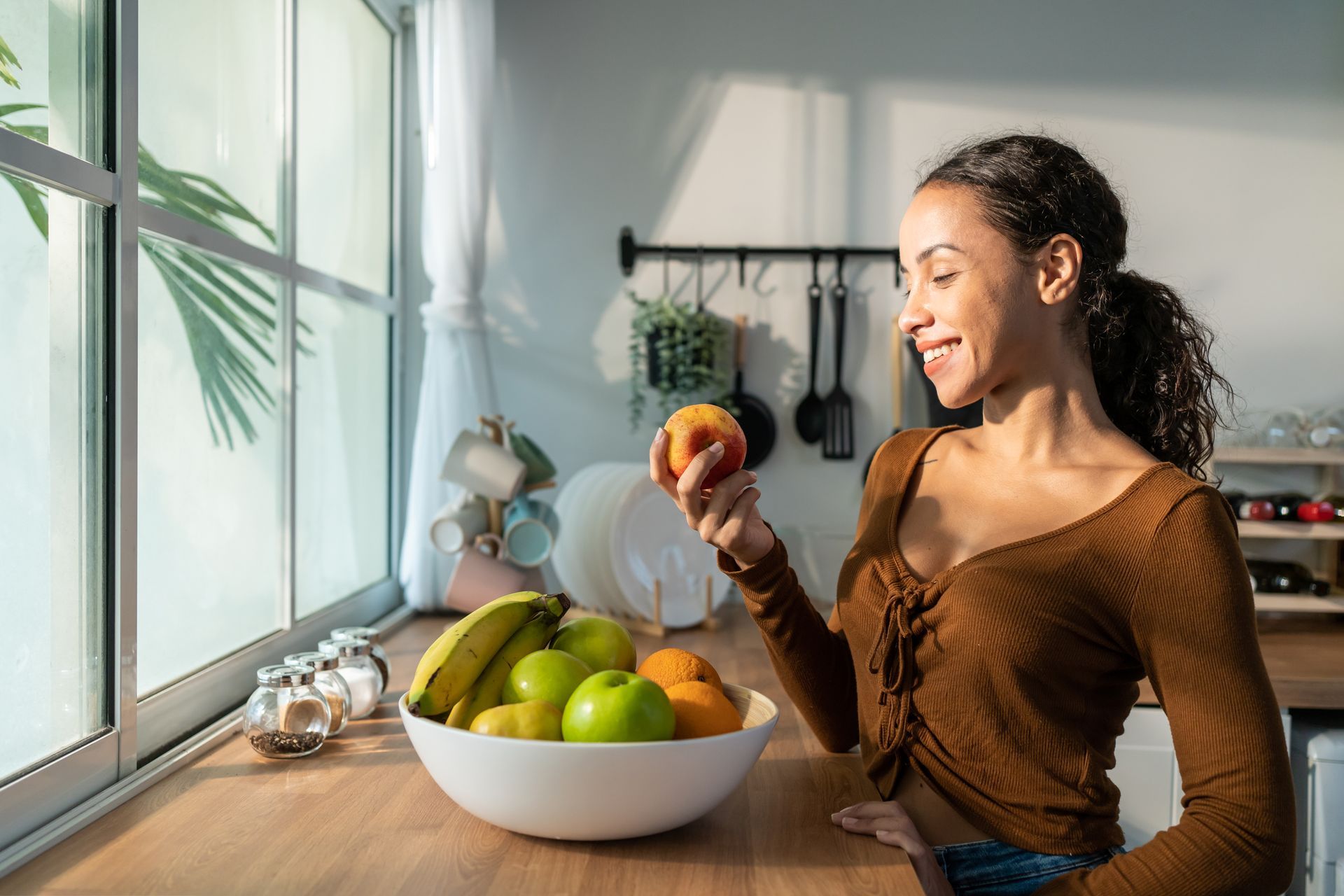 Woman in kitchen holding peach, smiling. Fruit bowl on counter by window.