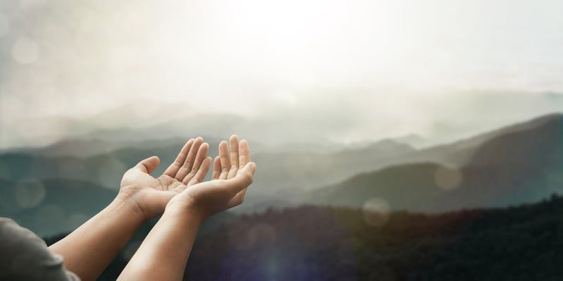 Hands cupped upwards, silhouetted against a mountain range and bright sky.
