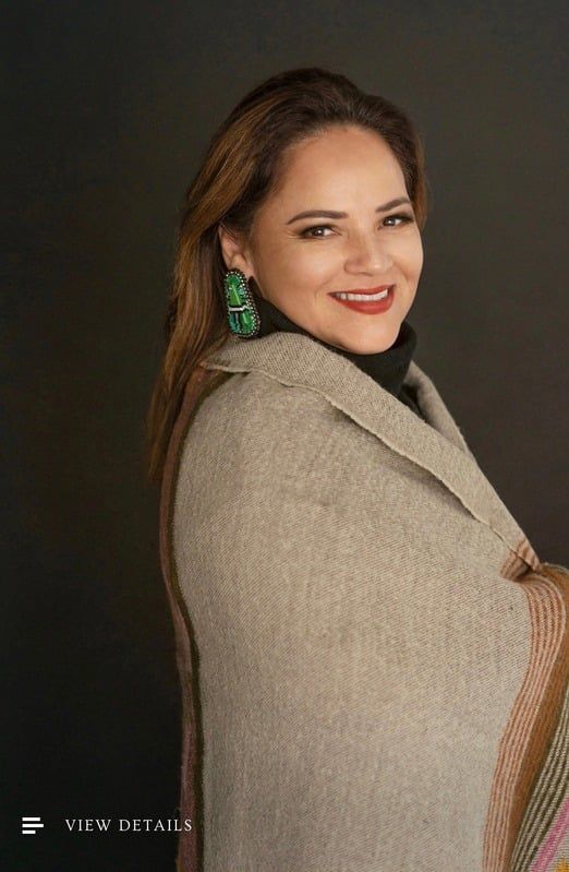 Woman with a smile, wearing a shawl and large green earrings, posed against a dark background.
