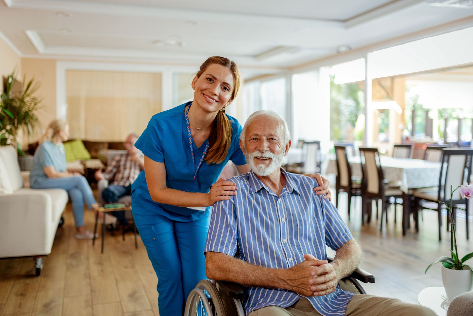 Nurse with arm around smiling elderly man in wheelchair; interior of a care facility.