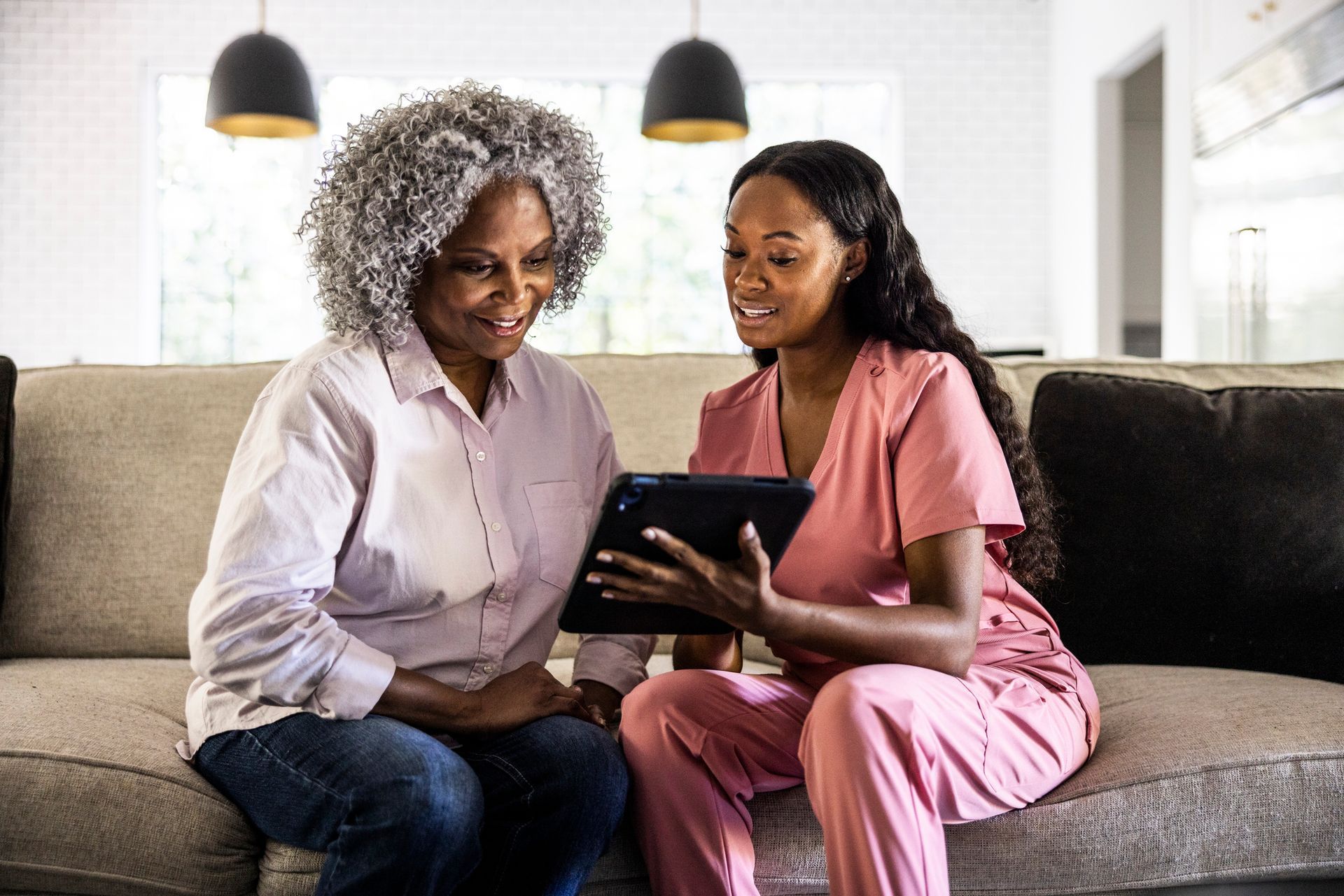 Woman and nurse looking at tablet on a couch. Nurse in pink scrubs, woman in light shirt and jeans.