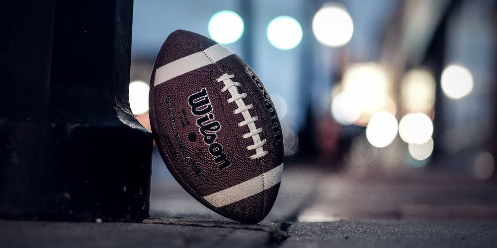 American football next to a black pole, on a sidewalk, blurred city lights in background.