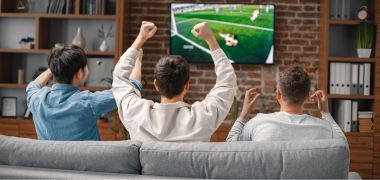 Three men are sitting on a couch watching a soccer game on a television.