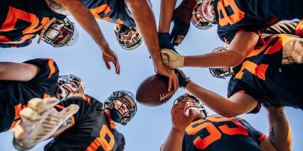 Football team huddles together, arms reaching for the football at the center. They wear navy and orange uniforms under a blue sky.