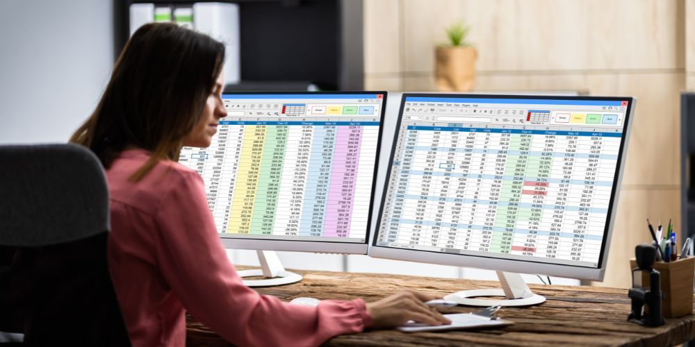 Woman working on spreadsheets displayed on dual computer monitors.