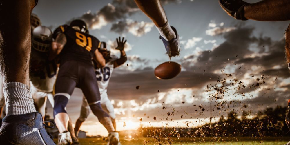 Football players in action on a field, a brown football in mid-air. A cloudy sky and setting sun in the background.