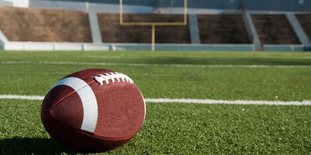 Football on a green field, with stadium seating and goalpost in the background.