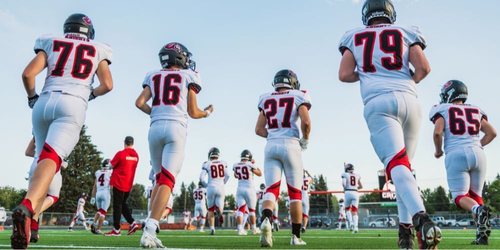Football players in white and red uniforms running on a green field towards the sun.