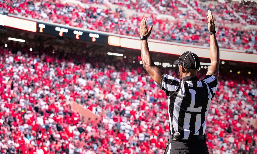 Football referee signals a touchdown with arms raised above a crowd of red and white fans in a stadium.