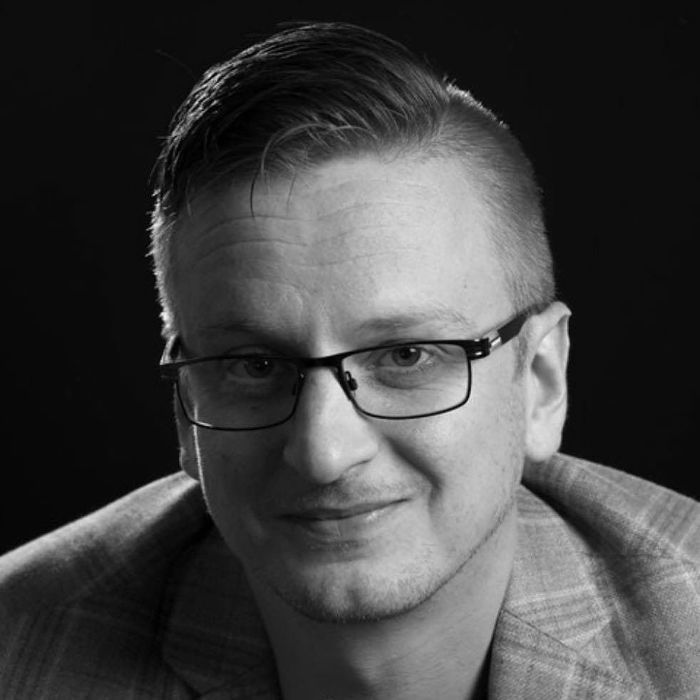 A man wearing glasses and a suit is smiling in a black and white photo.