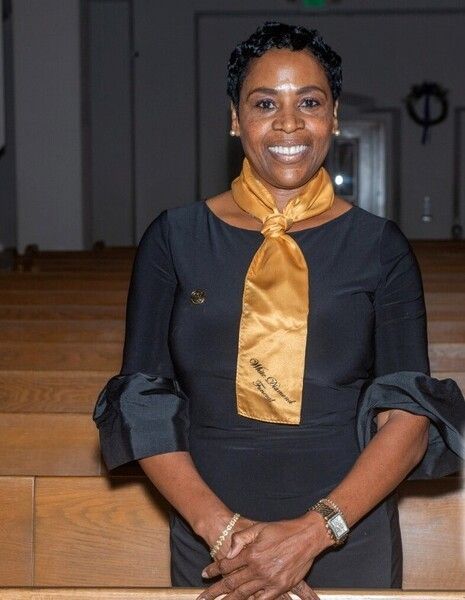 Woman in black dress with gold scarf, smiling in a church setting.