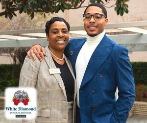 Woman in suit and man in blue jacket posing outdoors; White Diamond Funeral Services logo in corner.